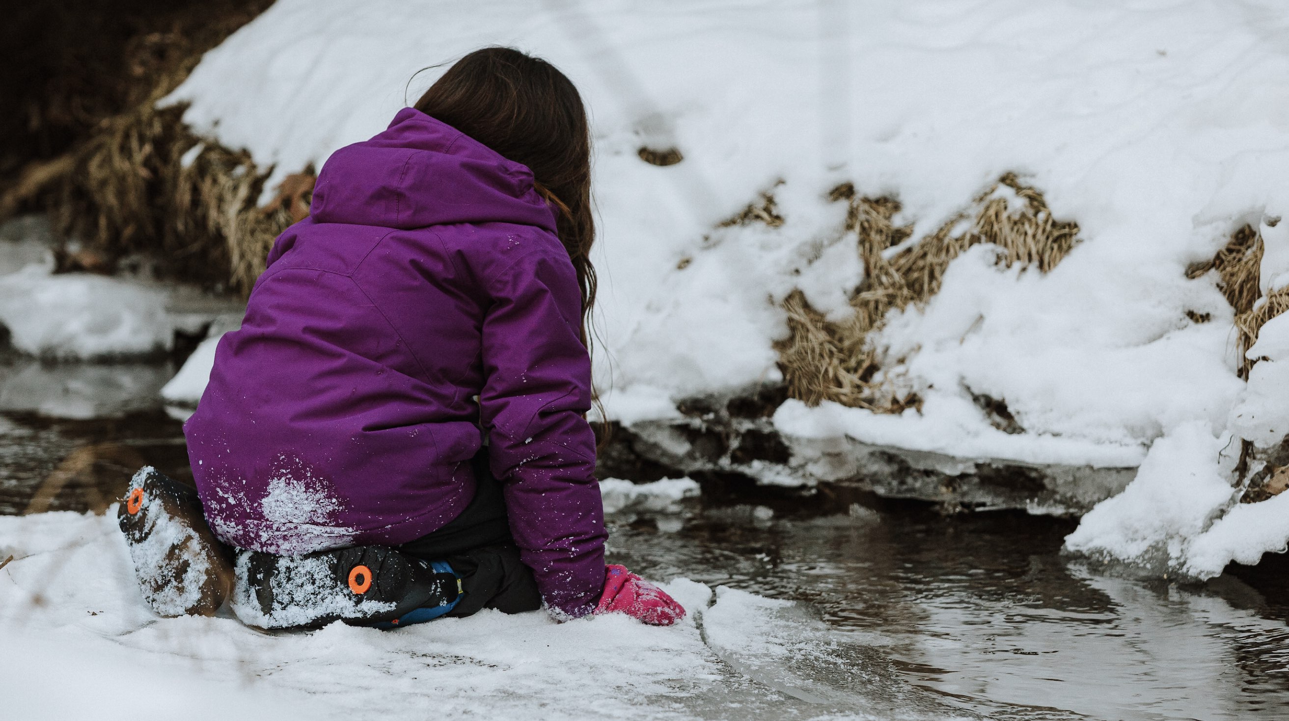 Image features a child kneeling by a frozen creek.