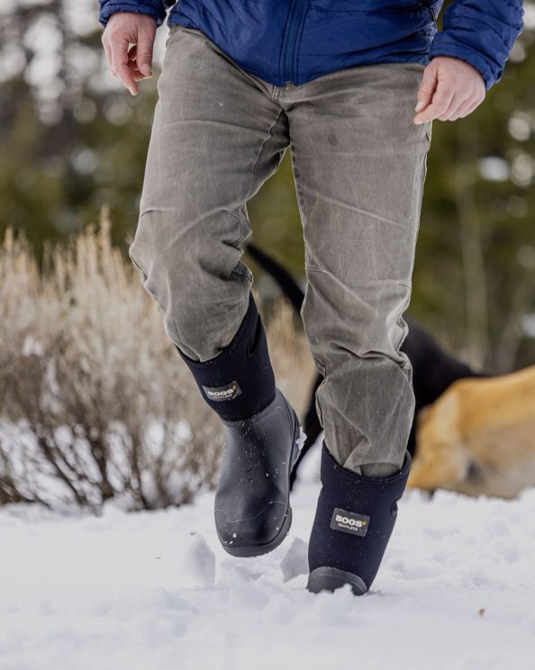 The image shown is of the Men's Bozeman tall insulated winter boots. The man is walking through the snow with his dogs in the background.