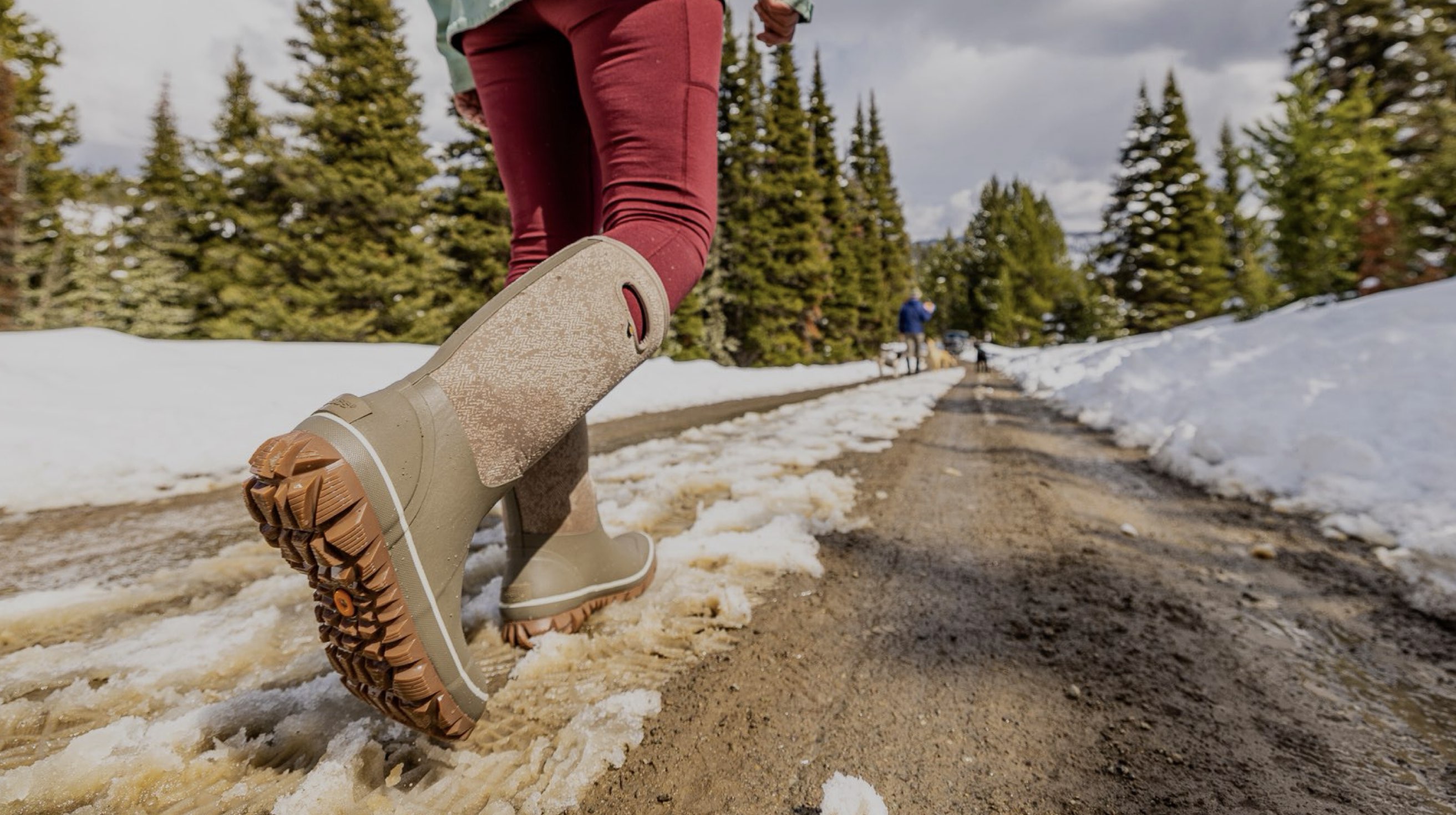 The image shown is of the Whiteout Faded tall insulated winter boots in a tan print. The woman is walking through the snow with his dogs in the background.