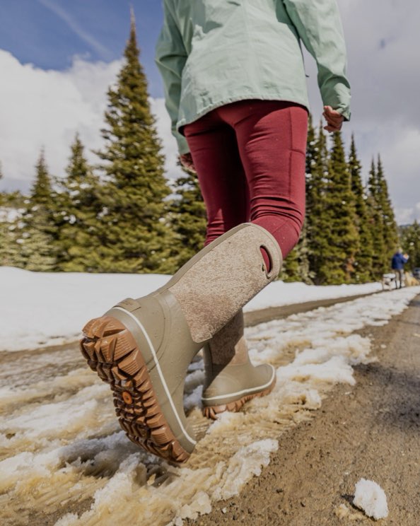 The image shown is of the Whiteout Faded tall insulated winter boots in a tan print. The woman is walking through the snow with his dogs in the background.