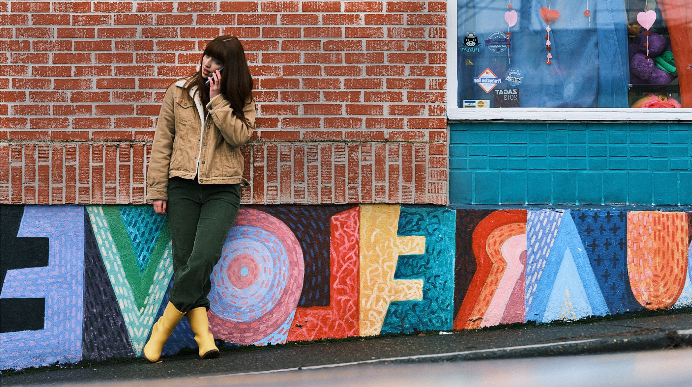 Shop our Women's New Arrivals.  The image shown is a woman leaning back on a brick wall, talking on the phone.  The boot highlighted our Women's Amanda Mid rainboots in yellow.