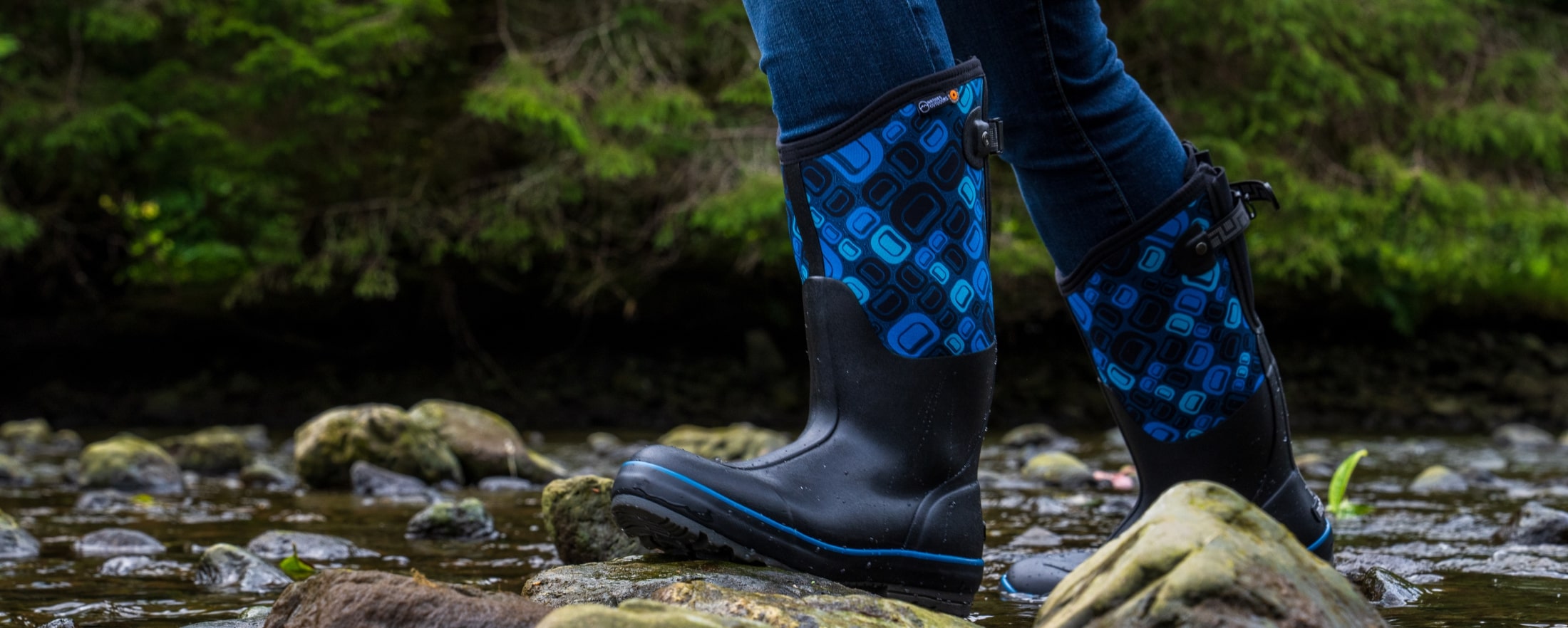 BOGS recently collaborated with Natives Outdoors and Tlingit jeweler Jennifer Younger to release a special-edition model of our Classic Il Adjustable Calf boot using her Tlingit formline ovoid design in various blue colors. The image shown is our Women's Classic II Adjustable calf Natives Outdoors boot walking through a river bed.