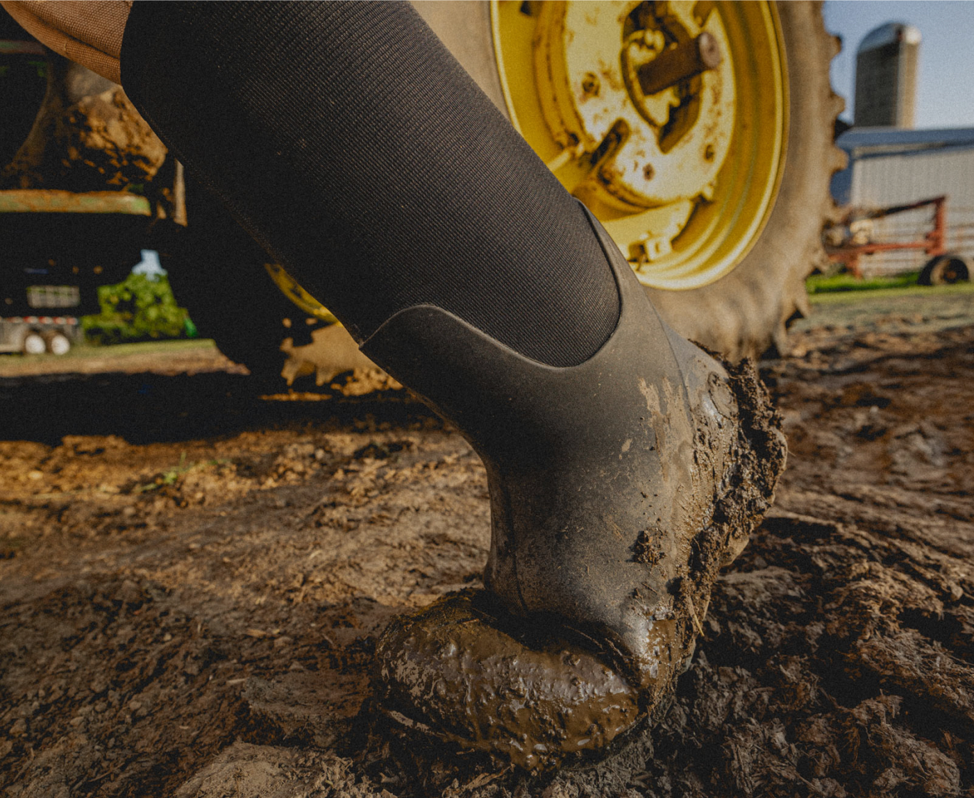 This is a closeup image of our Men's Classic Seamless boot tall walking through mud with a tractor in the background.