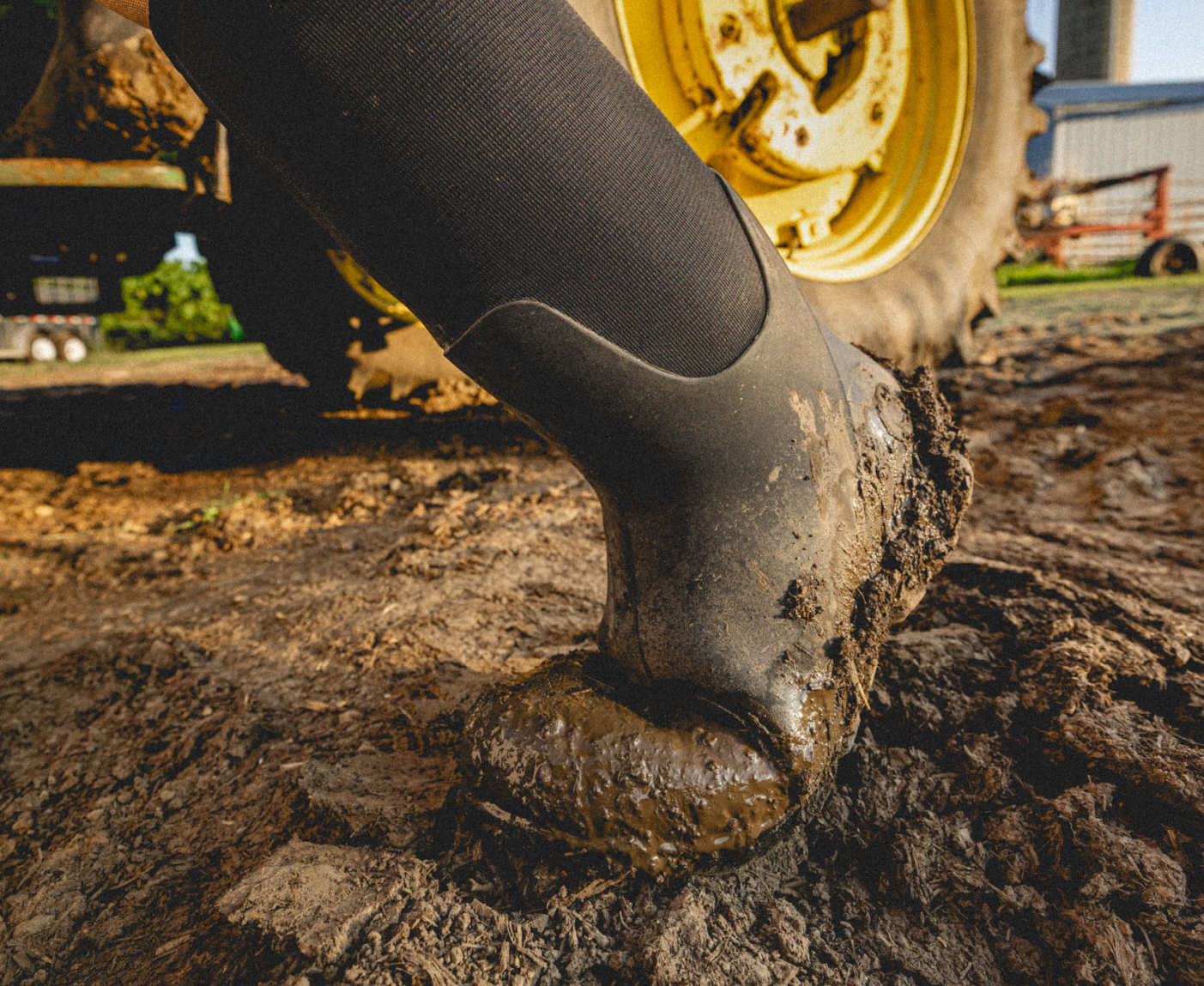 The image shown is a closeup image of our Men's Classic Seamless boot tall walking through mud with a tractor in the background.