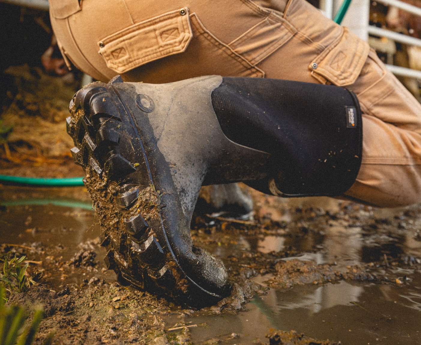 The image shown is a closeup image of our Men's Classic Seamless boot tall walking through mud with a tractor in the background.