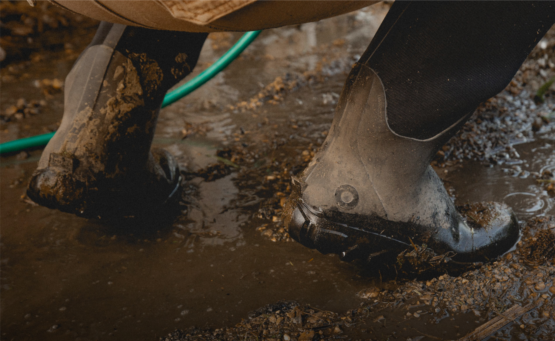 The image shown is a closeup image of our Men's Classic Seamless boot tall squatting down in muddy water with a hose in the background.