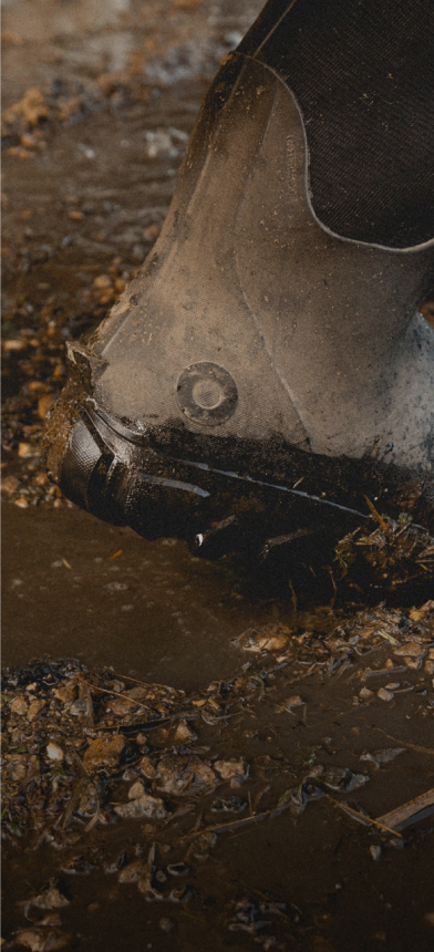 The image shown is a closeup image of our Men's Classic Seamless boot tall squatting down in muddy water with a hose in the background.