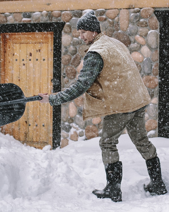 The image shown is a man shoveling snow outside of their cabin. Shop our top Men’s winter boots.