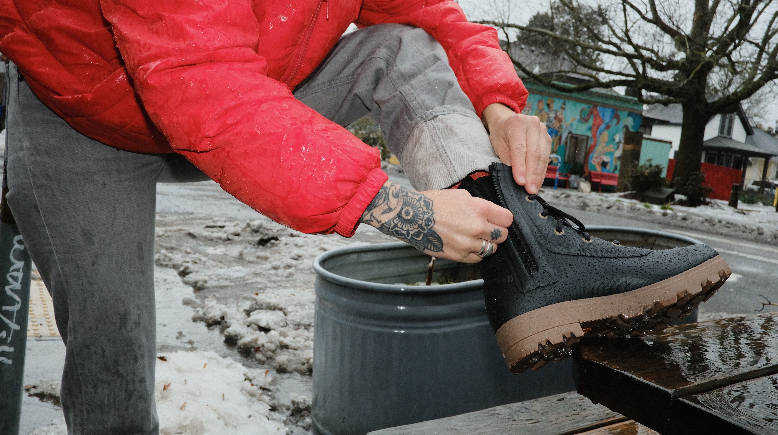 The image shown is a woman in a red jacket zipping up her Holly Rain Lace boots in black. The backdrop is on a winter urban street.