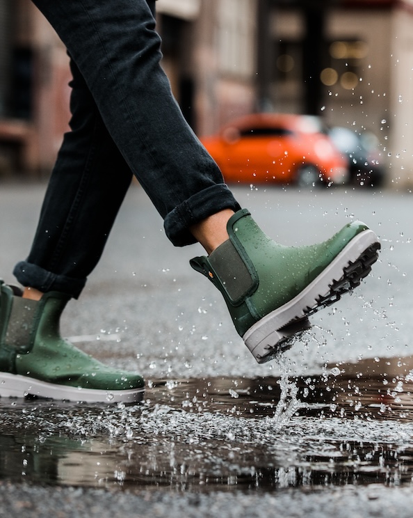 The image shown is a closeup of our women’s holly rainboot Chelsea in green splashing through a puddle. Shop now.